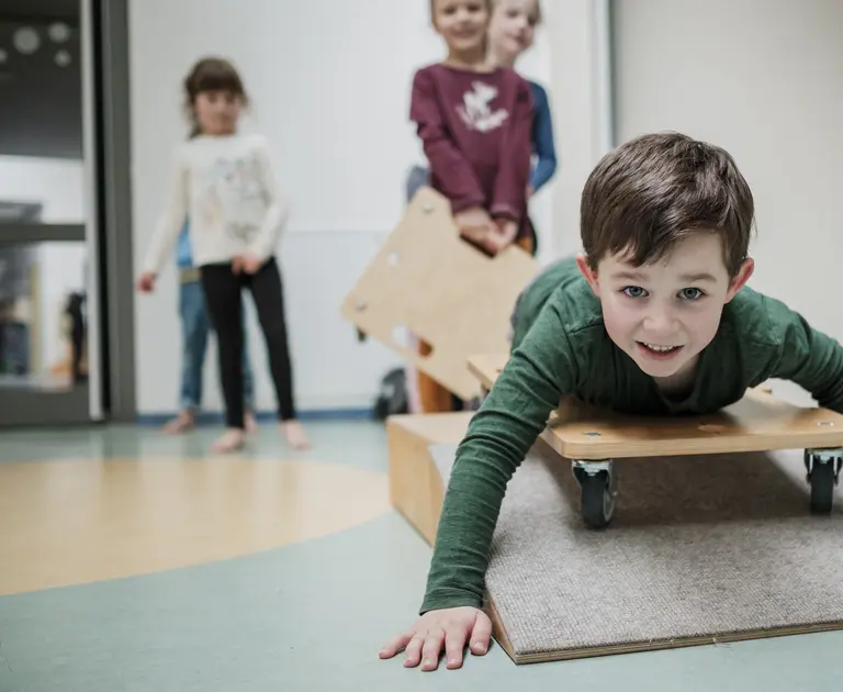 Junge liegt lachend auf einem Rollbrett. Im Hintergrund warten weitere Kinder auf ihren Einsatz. Das Bild vermittelt spielerische Bewegung, Gemeinschaft und Freude an inklusiver Förderung im CJD.