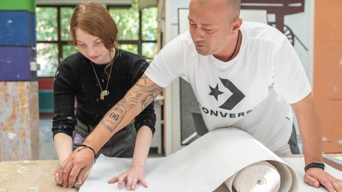 Erwachsener unterstützt Jugendliche beim Zuschneiden von Tapete im CJD, handwerkliches Lernen und individuelle Förderung.