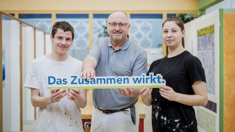 Ein Ausbilder für den Beruf Maler hält zusammen mit einem Auszubildenden und einer Auszubildenden ein Schild hoch mit dem Slogan des CJD "Das Zusammen wirkt".