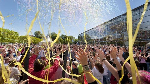 Große Menschenmenge feiert bei Sonnenschein mit bunten Bändern vor einem modernen Gebäude, Gemeinschaft und Inklusion beim CJD-Event.