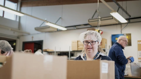 Eine Frau mit Down-Syndrom arbeitet konzentriert in einer Werkstatt und trägt dabei eine Haube. Im Hintergrund sind weitere Mitarbeitende zu sehen. Bei uns erhält jeder Mensch die Chance auf sinnvolle Arbeit und Teilhabe am Berufsleben. Entdecke unsere inklusiven Arbeitsangebote – jetzt informieren und neue Möglichkeiten schaffen!