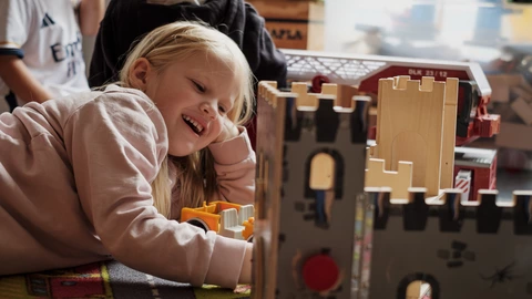 Ein blondes Mädchen liegt lachend auf dem Boden und spielt mit einer Ritterburg aus Holz. Um sie herum sind weitere Spielzeuge zu sehen, darunter ein Feuerwehrauto.