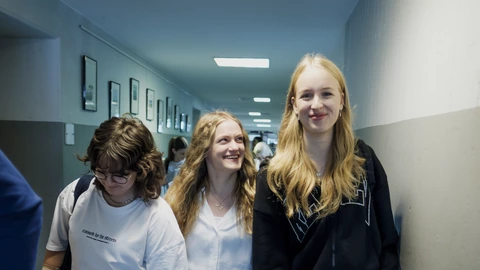 Drei Jugendliche gehen lachend und plaudernd durch einen hellen Schulflur mit grauen Wänden und gerahmten Bildern.