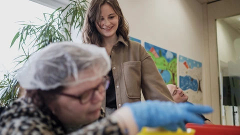 Eine junge Frau begleitet eine Frau mit Behinderung bei ihrer Arbeit in einer Werkstatt des CJD für Menschen mit Behinderung.
