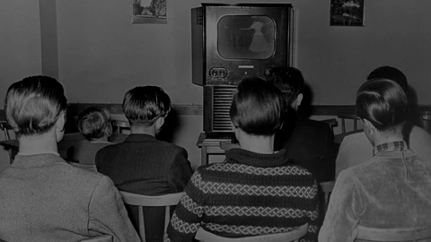7 Jugendliche im Alter von 14 -20 Jahren sitzen,mit dem Rücken zur fotografierenden Person, bei einer Freizeit in Dortmund vor einem Fernseher 1951. Auf dem Bildschirm ist das schwache Bild einer Frau im Kleid zu sehen. Das Bild ist in schwarz/weis dargestellt.  