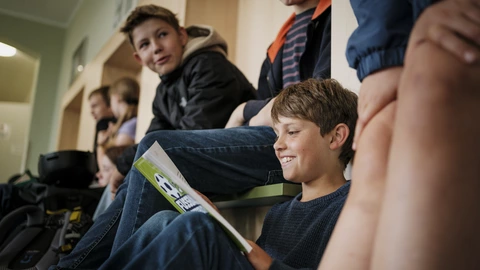 Junge liest lächelnd in einer Schulumkleide beim CJD, gemeinsames Lernen und Bildung in entspannter Atmosphäre.