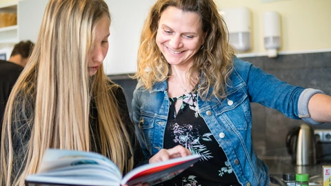 Eine Jugendliche steht neben einer Pädagogin am Herd. Sie schauen beide in ein Kochbuch. 