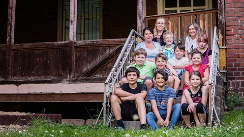 Eine Kindergruppe sitzt mit drei Pädagoginnen auf einer Treppe vor einem alten Gebäude.