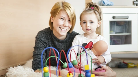 Eine junge Frau sitzt mit einem kleinen Mädchen auf dem Boden einer Kita. Das Kind hält eine Puppe im Arm, vor ihnen steht ein Holzspielzeug mit bunten Kugeln. Die Frau lächelt, das Kind blickt ruhig in die Kamera.