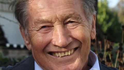 Farbfotografie eines älteren Mannes mit freundlichem Lächeln. Er trägt ein hellblau kariertes Hemd und eine dunkle Jacke. Der Mann hat graues Haar, Falten im Gesicht und wirkt vital. Im Hintergrund sind unscharf herbstliche Bäume und ein Gartenzaun zu sehen.