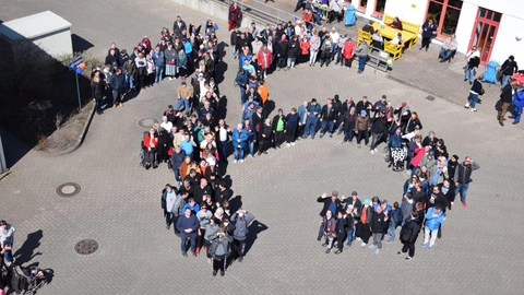 Drohnenaufnahme einer großen Menschengruppe, die auf einem gepflasterten Platz in Form einer großen „75“ steht. Die Personen sind bunt gekleidet, viele lächeln und winken in die Kamera. Im Hintergrund befindet sich ein Gebäude mit roten Fensterrahmen, gelben Sitzbänken und einigen weiteren Personen, die dem Geschehen zusehen. Die Szene findet bei sonnigem Wetter statt.