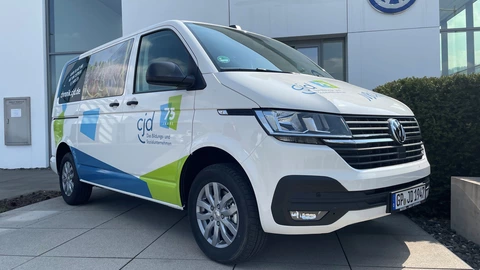 Weißer VW-Transporter mit farbigen CJD-Design-Elementen in Blau und Grün, seitlich mit dem Logo „75 Jahre CJD – Das Bildungs- und Sozialunternehmen“ und einer Fotografie beklebt. Der Wagen steht vor einem Volkswagen-Autohaus mit großem VW-Logo an der Fassade. Die Szene zeigt eine Fahrzeugübergabe bei sonnigem Wetter.