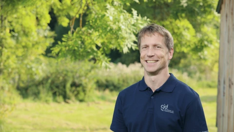 Ein Mann mit kurzen, hellbraunen Haaren steht lächelnd im Freien vor grüner, sommerlicher Naturkulisse. Er trägt ein dunkelblaues Poloshirt mit dem weißen CJD-Logo und dem Schriftzug „Das Bildungs- und Sozialunternehmen“. Im Hintergrund sind Bäume, Wiese und unscharfe Pflanzen zu sehen, die eine freundliche und natürliche Atmosphäre schaffen.