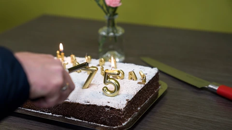 Ein rechteckiger Schokoladenkuchen mit Puderzucker ist mit goldenen Kerzen in Form der Zahl „75“ und weiteren Buchstaben dekoriert. Eine Hand entzündet die Kerzen mit einem Stabfeuerzeug. Daneben liegt ein großes Messer mit rotem Griff auf dem Tisch. Im Hintergrund steht eine kleine Glasvase mit einer rosa Blume vor grünlicher Wand.
