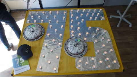 Auf einem gelben Tisch ist die Zahl 75 mit Alufolie ausgelegt und von vielen kleinen Teelichtern umrahmt. Zwei Kuchen mit Geburtstagskerzen in Form einer „7“ und „5“ stehen auf der Fläche. Daneben liegen eine schwarze CJD-Kappe und ein Flyer mit der Aufschrift „75 Jahre“. Die Dekoration erinnert an eine Feier zum Jubiläum. Im Hintergrund sind Teile eines Bildschirms und Möbel zu erkennen.