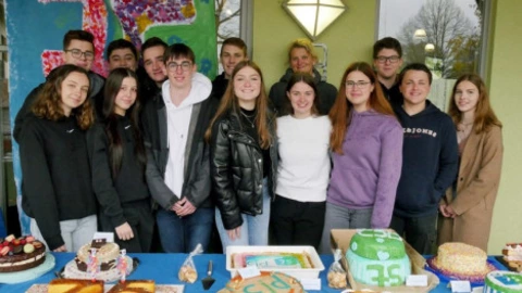 Eine Gruppe Jugendlicher steht dicht zusammen hinter einem Tisch mit vielen selbstgebackenen Kuchen. Auf dem Tisch liegen bunt verzierte Torten, darunter eine mit der Aufschrift „75“ und eine mit dem CJD-Logo. Im Hintergrund hängt ein farbig gestaltetes Plakat mit einer großen „75“. Alle lächeln in die Kamera, die Szene wirkt fröhlich und festlich.