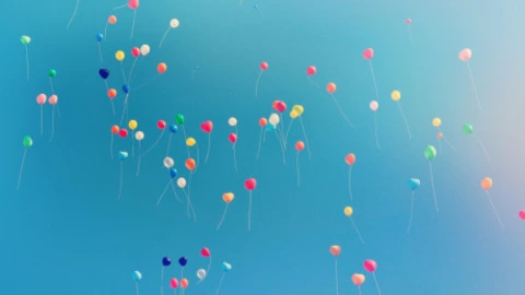 Vor strahlend blauem Himmel steigen zahlreiche bunte Luftballons in die Höhe. Die Ballons sind in verschiedenen Farben – Rot, Gelb, Blau, Grün, Rosa und Orange – und mit langen weißen Schnüren versehen. Sie schweben frei verteilt im Bild und erzeugen eine fröhliche, leichte und festliche Stimmung.