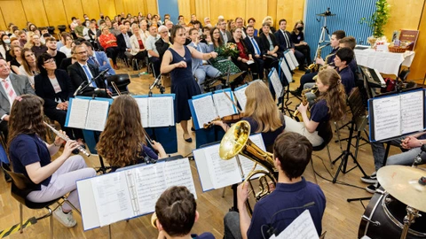 Eine Schülerband mit Blasinstrumenten und Schlagzeug spielt in einem Saal vor großem Publikum. Die Dirigentin steht in der Mitte und leitet das Ensemble. Im Hintergrund sitzen zahlreiche Gäste in feierlicher Kleidung, aufmerksam den jungen Musikerinnen und Musikern lauschend. Die Szene vermittelt eine festliche, lebendige Atmosphäre voller Musik und Gemeinschaft.