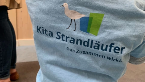 Ein kleines Kind trägt ein hellblaues T-Shirt mit dem Aufdruck „Kita Strandläufer – Das Zusammen wirkt.“ sowie einer Möwe als Logo. Das Kind steht mit dem Rücken zur Kamera, wodurch der Schriftzug und das Logo gut sichtbar sind. Im Hintergrund sind Erwachsene im Eingangsbereich der Einrichtung erkennbar. Die Szene vermittelt den Eindruck von Zugehörigkeit und Gemeinschaft in der Kindertagesstätte.