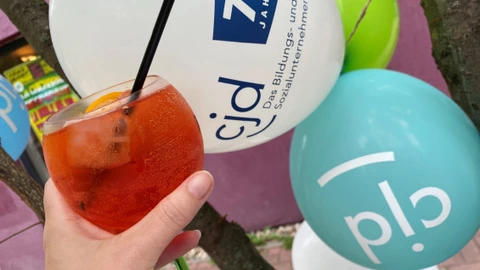 Eine Hand hält ein Glas mit einem Aperitif-Getränk, garniert mit Orangenscheiben und Strohhalm. Im Hintergrund sind bunte Luftballons mit dem CJD-Logo und dem Schriftzug „75 Jahre“ zu sehen. Die Szene vermittelt sommerliche Feierlaune und markiert den Anlass des 75-jährigen Jubiläums des CJD.