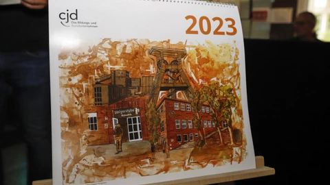 Ein Kalender des CJD für das Jahr 2023 steht auf einer Staffelei. Auf dem Titelblatt ist eine künstlerische Illustration eines Bergwerksgebäudes mit Fördertürmen und rotem Backstein zu sehen. Oben links steht das CJD-Logo mit dem Schriftzug „Das Bildungs- und Sozialunternehmen“, oben rechts die Jahreszahl „2023“ in Orange.