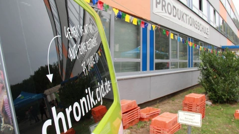 Ein grünes Auto mit der Aufschrift „chronik.cjd.de – Gehen Sie mit uns auf Entdeckungsreise. 75 Jahre CJD“ steht vor einem Gebäude der Produktionsschule. Am Gebäude hängt bunte Wimpel-Dekoration, und auf dem Hof sind gestapelte rote Kunststoffkisten zu sehen.