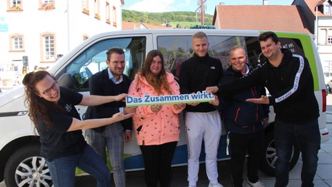 Sechs Personen stehen lachend vor einem CJD-Bus und halten gemeinsam ein Schild mit der Aufschrift „Das Zusammen wirkt.“ in die Kamera. Die Gruppe besteht aus jungen Erwachsenen und Mitarbeitenden, die eng nebeneinanderstehen und den Teamgeist symbolisieren. Im Hintergrund sind Häuser und ein sonniger Himmel zu sehen.