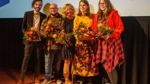 Fünf Personen stehen nebeneinander auf einer Bühne vor einer Leinwand und halten Blumensträuße in den Händen. Sie lächeln in die Kamera, eine Person trägt zusätzlich ein Mikrofon. Die Gruppe wirkt feierlich und harmonisch, als sei es der Abschluss oder die Ehrung bei einer Veranstaltung, etwa einer Filmpremiere oder Preisverleihung.