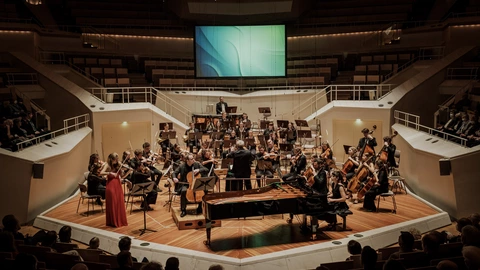 Ein Orchester spielt in einem großen Konzertsaal vor Publikum. Im Vordergrund ist ein Flügel sichtbar, daneben eine Solistin im roten Kleid an der Violine. Der Dirigent steht in der Mitte vor den Musikerinnen und Musikern. Die Bühne ist hell ausgeleuchtet, im Hintergrund hängt eine große Leinwand. Die Atmosphäre wirkt festlich und konzentriert.