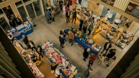 Blick von oben auf einen weihnachtlich dekorierten Markt in einer Halle. Menschen schlendern zwischen Ständen mit bunten Handarbeiten, Dekorationen und Geschenkartikeln. Einige Besucher unterhalten sich, andere betrachten die Auslagen. Festliche Beleuchtung und geschmückte Tische sorgen für eine warme, gemütliche Atmosphäre.