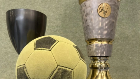 Ein gelb-grüner Softfußball liegt vor zwei Pokalen. Der rechte Pokal ist groß, silbern mit goldenen Akzenten und trägt die Gravur „CJD Dortmund Soccercup Wanderpokal“. Links daneben steht ein kleinerer, dunkler Pokal. Das Bild vermittelt eine sportliche Atmosphäre und erinnert an ein Fußballturnier mit Siegerauszeichnung.