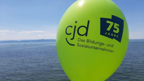 Ein grüner Luftballon mit dem blauen Aufdruck „cjd – Das Bildungs- und Sozialunternehmen“ sowie dem Logo „75 Jahre“ schwebt vor blauem Himmel am Ufer eines Sees. Das Bild symbolisiert das Jubiläum des CJD in festlicher, sommerlicher Atmosphäre.