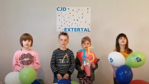 Vier Kinder stehen nebeneinander vor einer Wand mit einem bunten Plakat „CJD Extertal“. Sie halten Luftballons in Blau, Grün und Weiß mit CJD-Logo sowie eine bunte „75“-Zahl in den Händen. Die Szene vermittelt Freude und Zusammenhalt und zeigt eine Aktion zum 75-jährigen Jubiläum des CJD.