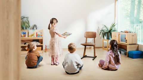 Drei Kinder sitzen auf dem Boden einer CJD Kita und schauen auf einen leeren Stuhl für Erwachsene. Ein Mädchen steht und reicht ein Buch zum leeren Stuhl, als würde dort Jemand sitzen.