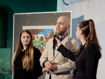  Stephan Zwingmann spricht bei der Ausstellung im Rahmen des CJD Orchesterkonzerts 2023 im Foyer der Berliner Philharmonie in ein Mikrofon, das von einer jungen Frau rechts neben ihm gehalten wird. Links neben ihm steht eine weitere junge Frau. Im Hintergrund hängt ein Bild aus der Ausstellung.