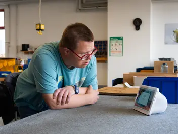 Ein Mensch lehnt mit verschränkten Armen an einem Tisch und schaut auf ein Alexa Gerät.