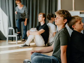 Vier Jugendliche sitzen Rücken an Rücken. Sie haben die Augen geschlossen. Im Hintergrund steht eine Frau.