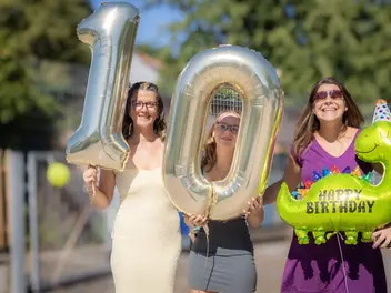 3 Personen halten große Heliumballons in den Händen. Zu sehen ist die Zahl 10 und ein grüner Dinosaurier mit der Aufschrift "Happy Birthday".