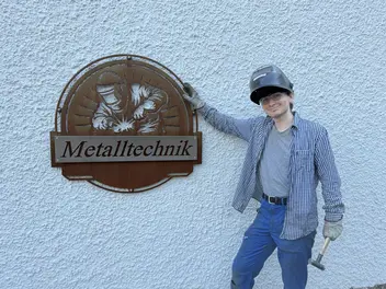 Maciej Lehr steht mit hochgeklappter Schweißmaske und Werkzeug in der linken Hand vor einer weißen Hauswand rechts neben einem runden Schild aus Metall auf dem Metalltechnik steht.