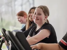 Eine Jugendliche macht Sport auf einem Fitnessgerät. Links und rechts von ihr sind weitere Jugendliche auf Fitnessgeräten zu sehen. Spenden helfen einen Fitnessraum in einer Jugendwohngruppe auszustatten.