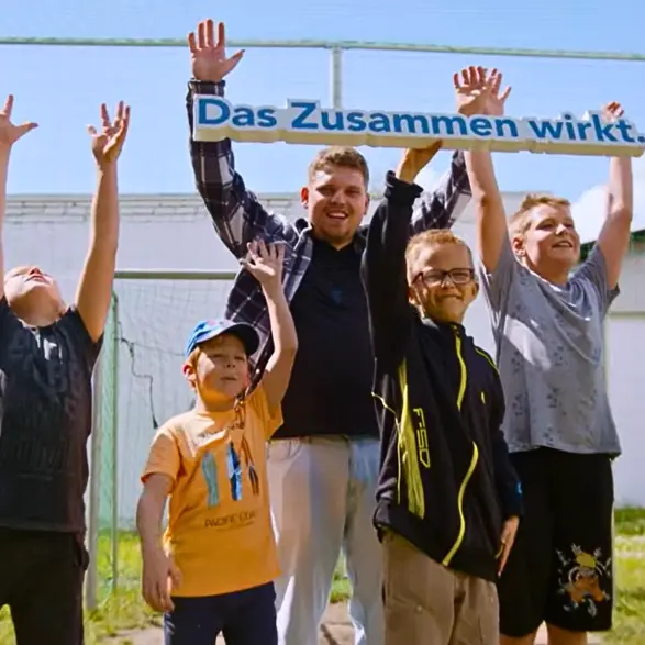 Mehrere Kinder und ein Erzieher des CJD jubeln und halten ein Schild mit dem CJD-Motto Das Zusammen wirkt