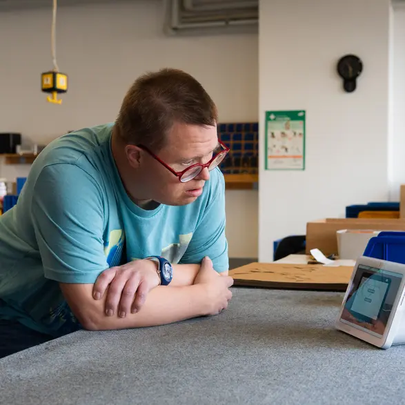 Ein Mensch lehnt mit verschränkten Armen an einem Tisch und schaut auf ein Alexa Gerät.