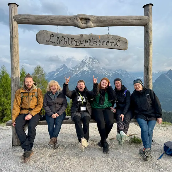 Sechs Personen auf einer Bank mit einem Schild "Lieblingsplatzerl". Im Hintergrund sind Berggipfel.