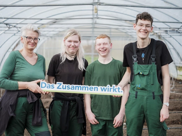 Vier Menschen in einem Gartenhaus halten ein Schild mit der Aufschrift "Das Zusammen wirkt"