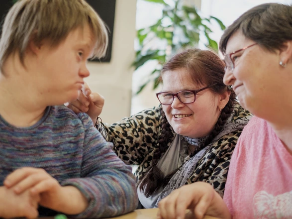 Drei Frauen, teilweise mit Behinderung, interagieren miteinander und lächeln sich an. Spenden ermöglichen eine fürsorgliche Betreuung in Wohngruppen für Menschen mit Behinderung.