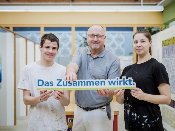 3 Leute halten zusammen den Slogan "Das Zusammen wirkt." und lächeln