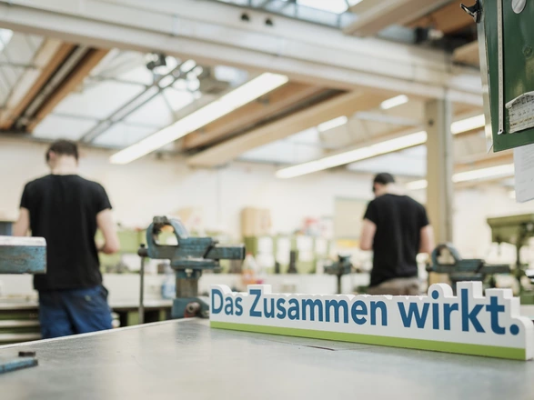 Schriftzug „Das Zusammen wirkt.“ in einer Werkstatt des CJD, zwei Menschen arbeiten im Hintergrund.