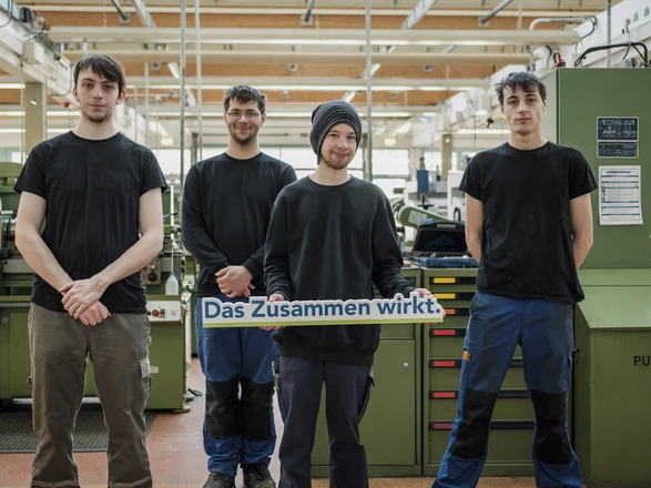 Vier junge Männer stehen nebeneinander lächelnd in einer Werkstatt vor Maschinen. Einer in der Mitte hält den Schriftzug "Das Zusammen wirkt.". Spenden helfen jungen Menschen in unseren Bildungsangeboten ihre Zukunft zu gestalten.