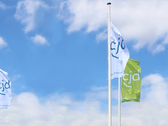 CJD-Fahnen in Blau, Weiß und Grün wehen vor blauem Himmel, Zeichen für Vielfalt und Gemeinschaft im CJD.