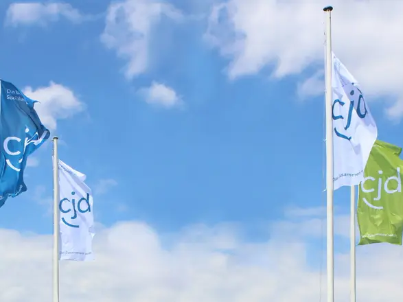 CJD-Fahnen in Blau, Weiß und Grün wehen vor blauem Himmel, Zeichen für Vielfalt und Gemeinschaft im CJD.